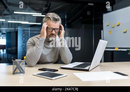 Mal di testa di tensione sul lavoro. Un giovane uomo d'affari con i capelli grigi negli occhiali siede in ufficio alla scrivania con un computer portatile e un telefono, sente un mal di testa, tiene la testa con le mani. Foto Stock