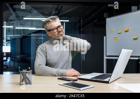 Un uomo d'affari stanco ed esausto in occhiali siede in ufficio alla scrivania, gli tiene il collo, gli massaggia il collo con la mano. Sente dolore nei suoi muscoli. Foto Stock