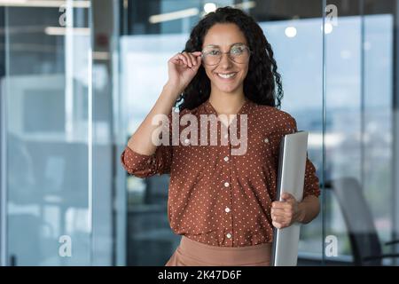 Ritratto di una giovane donna ispanica. Donna latinoamericana architetto e designer si trova in ufficio con progetti e disegni in mano, tiene gli occhiali in mano, guarda la fotocamera, sorride. Foto Stock