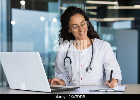 Formazione online. Giovane bella ragazza latino-americana studenti studi medici, prende l'esame online a distanza a casa dal computer portatile. Seduto al tavolo in un abito medico, scrivendo in un notebook. Foto Stock