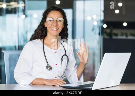 Ritratto di una giovane donna ispanica medico seduto in un abito medico con uno stetoscopio in ufficio. Funziona con i pazienti online con un notebook. Guarda la macchina fotografica, sorride, fa ondare la mano Foto Stock