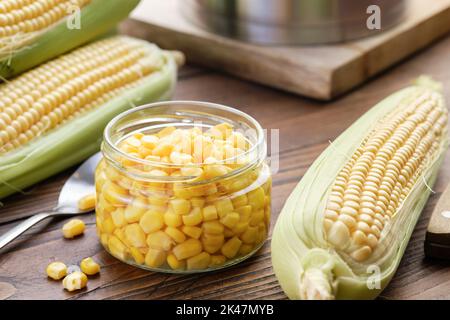 Granoturco dolce sottaceto in un vaso di vetro, cornocie fresche, pentola sul tavolo da cucina. Foto Stock