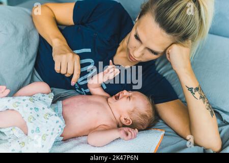 giovane mamma caucasica che si posa a letto con il suo bambino di due mesi. Suo figlio sta indossando un pannolino, urlando e piangendo e sta cercando di calmarlo. Foto di alta qualità Foto Stock