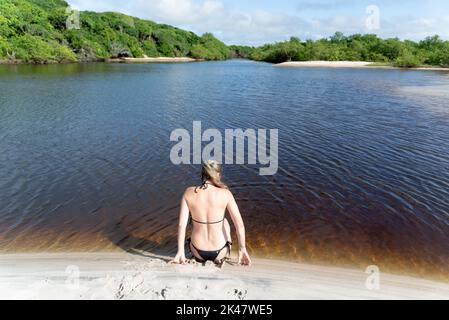 Una donna in un bikini seduta con la schiena alla macchina fotografica presso il fiume contro la foresta sullo sfondo. Guaibim spiaggia nella città di Valenca, Bahia Foto Stock