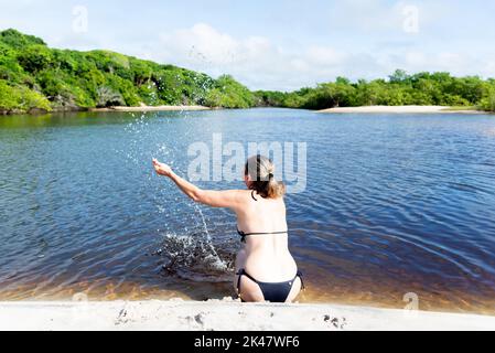 Donna in un bikini seduto sulla riva del fiume spruzza acqua verso l'alto contro la foresta e il cielo sullo sfondo. Guaibim spiaggia nella città di V Foto Stock