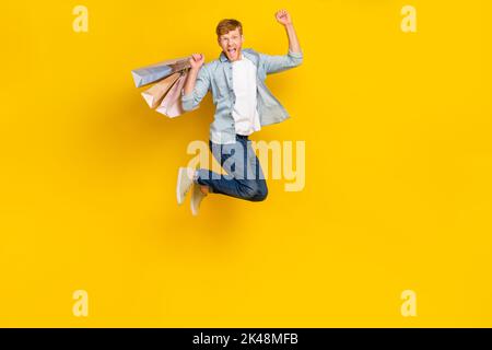 Foto a grandezza naturale del fortunato bel ragazzo rosso capelli indossare camicia blu jeans sneakers impressionato salto pugno in su isolato su sfondo di colore giallo Foto Stock