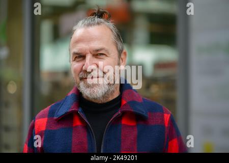 Uomo di mezza età con un sudicio mordente in una giacca in stile lumberjack rosso e blu si alza con un sorriso morbido in una strada commerciale Foto Stock