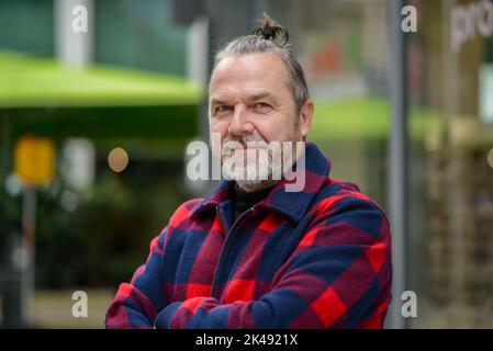 Uomo di mezza età con una mordente pistola in una giacca rossa e blu nello stile di un lumberjack si alza con le braccia incrociate in una strada commerciale Foto Stock