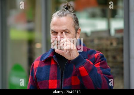 L'uomo di mezza età con un sudicio mazzetto in una giacca in stile lumberjack rosso e blu è in piedi in una strada commerciale e indica al suo occhio anteriore Foto Stock