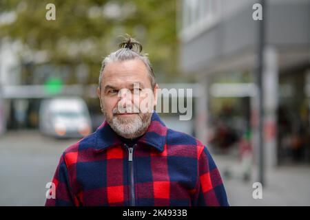 Uomo di mezza età con un sudicio muso in una giacca in stile lumberjack rosso e blu si erge con un aspetto disdicevole a lato in una strada commerciale Foto Stock