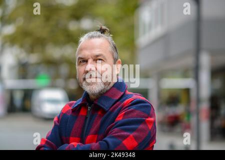 Uomo di mezza età con una mordente pistola in una giacca rossa e blu nello stile di un lumberjack si alza con le braccia incrociate in una strada commerciale Foto Stock