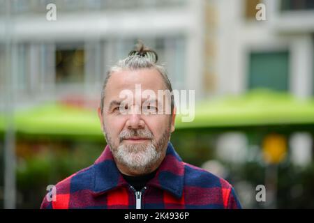 Uomo di mezza età con un sudicio mordente in una giacca in stile lumberjack rosso e blu si trova in una strada commerciale con un sorriso morbido Foto Stock