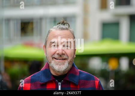 L'uomo di mezza età con un sudicio muso in una giacca in stile lumberjack rosso e blu si trova in una strada commerciale con un sorriso malizioso Foto Stock