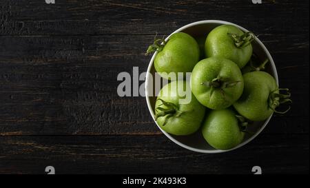 tazza bianca piena di pomodori, bacche verdi raccolte di fresco su un piano rustico in legno nero, preso dall'alto con spazio copia Foto Stock