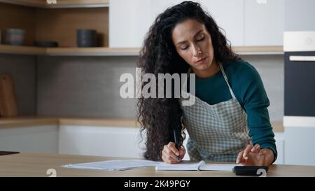 Donna ispanica concentrata moglie giovane mamma stand in cucina moderna prendere appunti a copybook scrivere ricetta elenco di alimentari calcolare l'uso di utenze domestiche Foto Stock