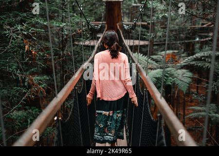 giovane donna caucasica piacevole passeggiando lungo una lunga e stretta passerella di legno che collega gli alberi della foresta lussureggiante, sequoia, rotorua, nuovo Foto Stock