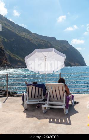 Una foto verticale di persone su sedie a sdraio sulla riva della città costiera di Porto Moniz Foto Stock