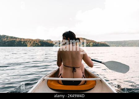 ragazza caucasica che indossa un costume da bagno seduto all'interno di un kayak sulla schiena e che guarda sul lato del lago in direzione dell'isola, il lago di tarawera Foto Stock