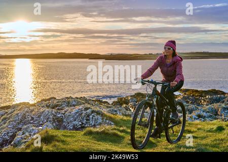 Bella donna anziana in mountain bike, in bicicletta al tramonto sulla spiaggia di sabbia dorata di Sillerna, Grallagh, County Galway, nella parte occidentale della Repubblica Foto Stock