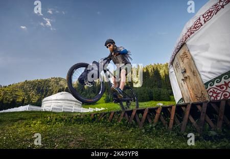 Uomo con greggi e la barba cavalcare e saltare in mountain bike con borse turistiche vicino Nomad Yurt casa nella bella valle di montagna in Almaty, Kazakhsta Foto Stock