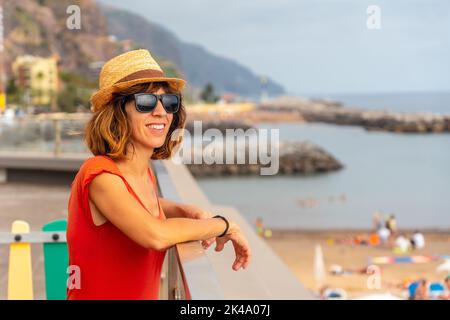 Un ritratto di una giovane donna turistica in abito rosso sulla spiaggia di Praia da Calheta in estate, Madeira. Foto Stock