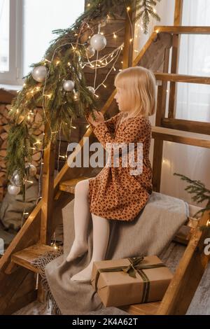 Una piccola ragazza bionda è seduta su una scala di legno in un interno scandinavo decorato in uno stile di Capodanno. Un bambino tiene un regalo di Natale in un Foto Stock