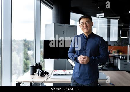 Programmatore in occhiali e camicia sorridente e guardando la telecamera, uomo asiatico che lavora all'interno di una moderna azienda informatica, che lavora in ufficio con diversi computer e monitor. Foto Stock