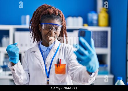 Donna africana che lavora al laboratorio scienziato facendo videochiamata urlando orgogliosa, celebrando la vittoria e il successo molto eccitato con braccio sollevato Foto Stock