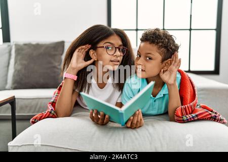 Due fratelli sdraiati sul divano leggendo un libro sorridendo con la mano sopra l'orecchio ascoltando una voce di voce o di pettegolezzo. Concetto di sordità. Foto Stock
