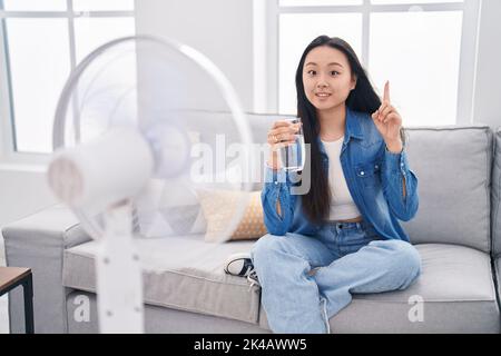 Giovane donna asiatica bere un bicchiere d'acqua godendo di aria dal ventilatore sorridendo con un'idea o domanda puntando il dito con il volto felice, numero uno Foto Stock