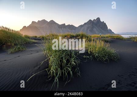 Turista su una duna di sabbia, spiaggia nera con sabbia vulcanica, spiaggia di sabbia, dune con erba, punta di Stokksnes, catena montuosa di Klatindur, Austurland Foto Stock