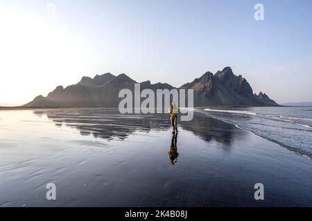 Turista alla spiaggia di lava nera, spiaggia sabbiosa e mare, montagne Clifatindur riflesso in acqua, Eystrahorn e Kambhorn, punta Stokksnes, montagna Foto Stock