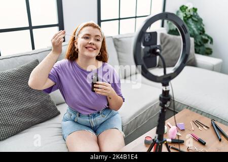 Giovane donna rossa che fa un tutorial sul trucco con smartphone a casa Foto Stock