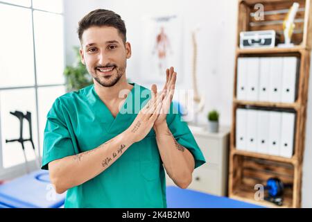 Giovane uomo fisioterapista che lavora in clinica di recupero del dolore che batte e applaudendo felici e gioiosi, sorridendo mani orgogliose insieme Foto Stock