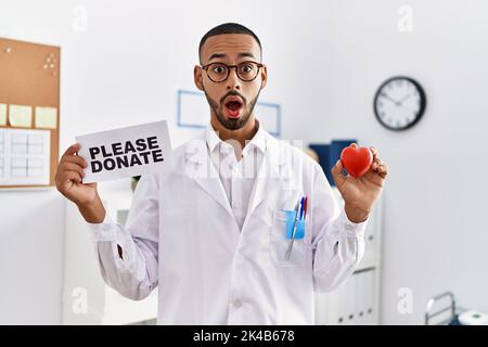 Dottore afro-americano che sostiene le donazioni di organi in faccia d'urto, guardando scettico e sarcastico, sorpreso con la bocca aperta Foto Stock