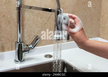 Una mano applica un detergente alla superficie di un rubinetto da cucina con una spugna Foto Stock