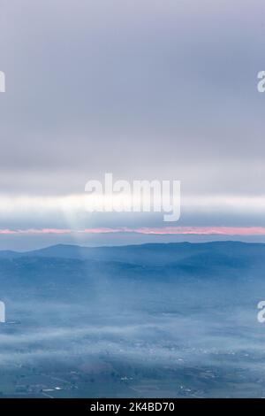 Raggi di sole che scende da alcune nuvole sopra una valle piena di nebbia, illuminando una parte di esso Foto Stock