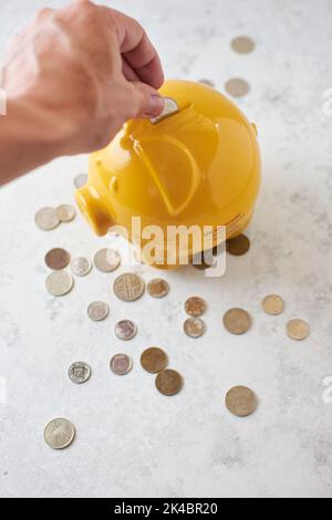 Una mano che mette le monete all'interno di un pigg giallo accanto alle monete su una superficie di marmo bianco Foto Stock