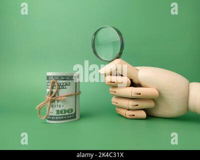 Una mano di legno che tiene la lente d'ingrandimento con le banconote su uno sfondo verde Foto Stock