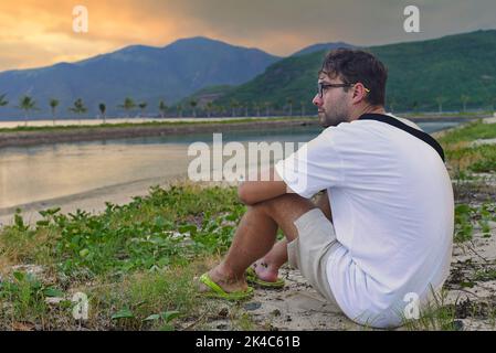 Uomo sconvolto seduto in spiaggia e guardando il tramonto nei tropici del Vietnam Foto Stock