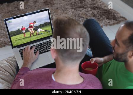 Due amici maschi diversi a casa che guardano una partita di rugby sul laptop. Immagine composita digitale di sport, competizione, intrattenimento e tecnologia. Foto Stock