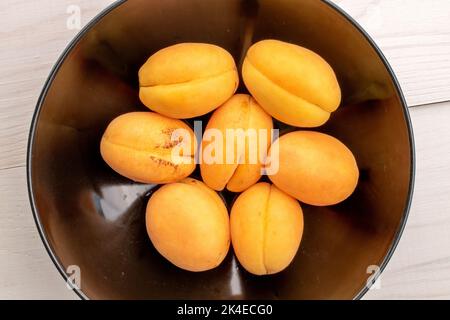 Diverse albicocche di ananas gialle mature in un piatto di ceramica nera, primo piano, su un tavolo di legno bianco, vista dall'alto. Foto Stock