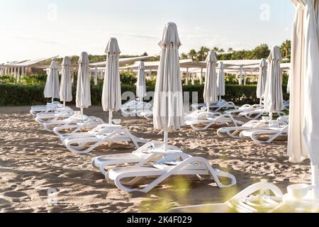 File di ombrelloni ripiegati e lettini vuoti sullo sfondo di sabbia e mare. Resort hotel. Concetto di fine stagione turistica. Foto Stock