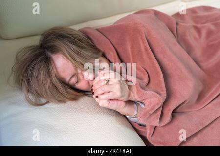 Donna sdraiata su un divano e che fa freddo durante l'inverno e il risparmio energetico, soffiando le mani per riscaldarsi. Foto Stock