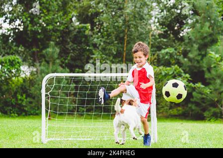 Il bambino eccitato che gioca il calcio calcia la palla mentre il suo cane salta per catturarla Foto Stock