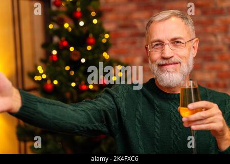 Un uomo anziano in una maglia verde che parla con la famiglia dal collegamento video del telefono vicino all'albero di Natale Foto Stock