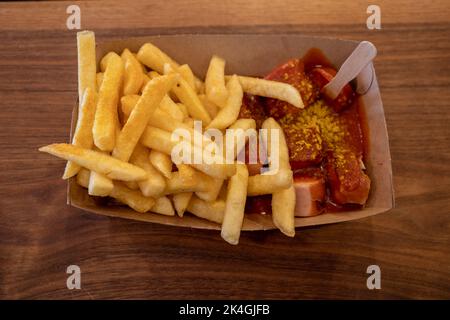 Vista dall'alto, Currywurst, salsiccia di maiale alla griglia in cima con curry in polvere servito con salsa al curry ketchup e patate fritte su vassoio di carta e tavolo di legno Foto Stock