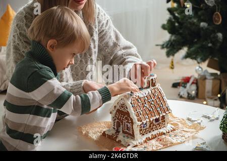 Ragazzino con madre che decora insieme casa di pan di zenzero di natale, attività di famiglia e tradizioni a Natale e Capodanno Foto Stock