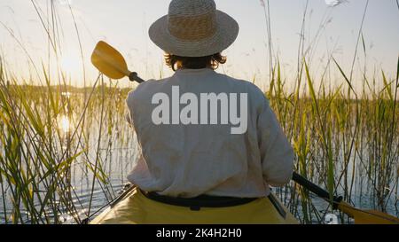 Persona che indossa vele con cappello di paglia in kayak utilizzando pagaia attraverso il rush in acqua. L'uomo turistico apprezza la canoa al tramonto vicino vista sul retro Foto Stock