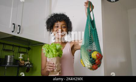 La donna afroamericana tiene lattuga e sacchetto della maglia con le verdure fresche. Casalinga con prodotti biologici stand in cucina sorridente Foto Stock